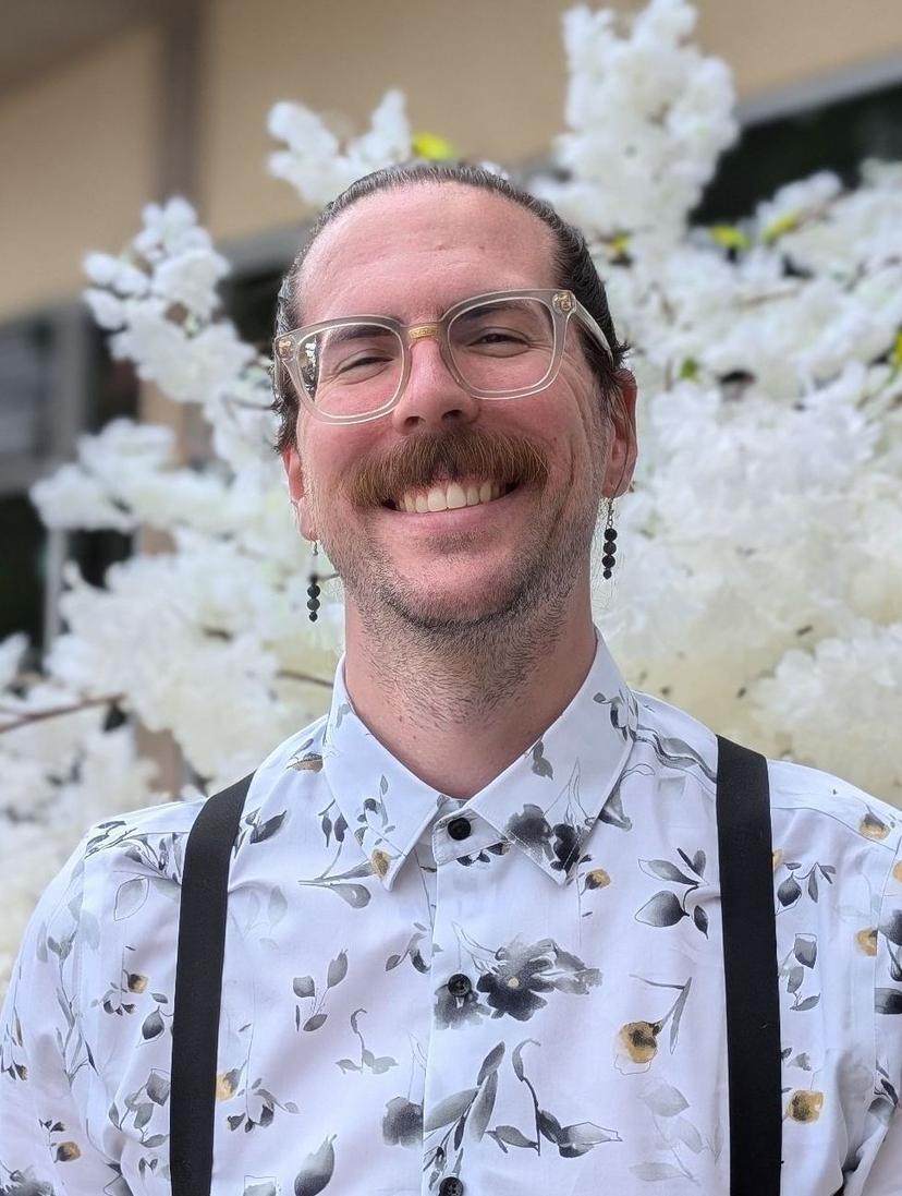 Picture of Mitchell Coovert. He is wearing a white floral dress shirt, and standing in front of a tree of off white flowers. 