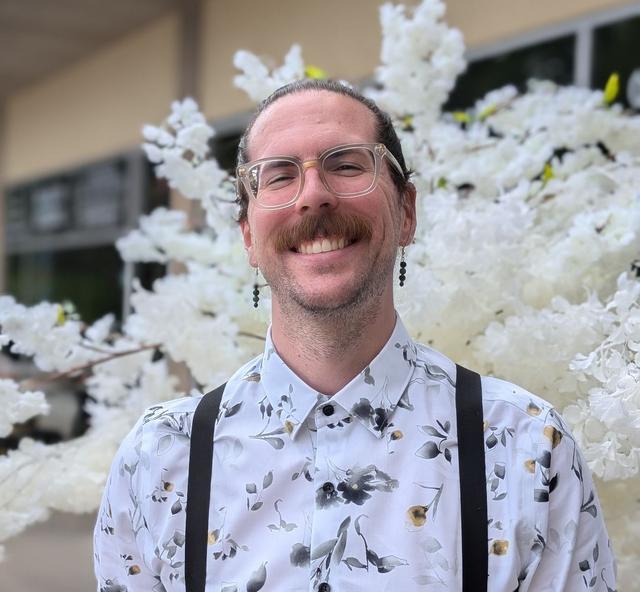 Picture of Mitchell Coovert. He is wearing a white floral dress shirt, and standing in front of atree of off white flowers.